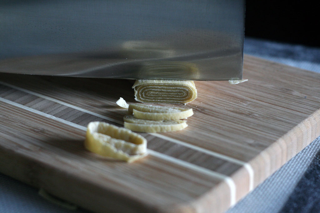Close-up of knife about to slice through rolled up ramen noodle dough. The rolled up ramen noodle dough has a spiral of flour.
