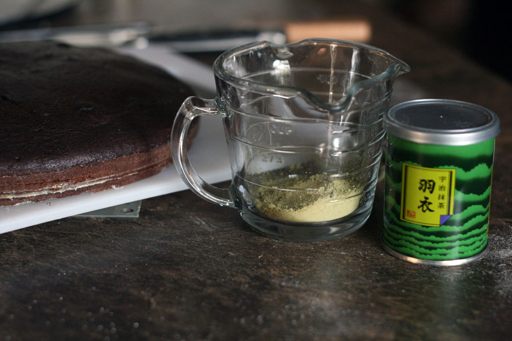 A glass measuring cup with some matcha green tea powder sitting beside a can of green tea powder.