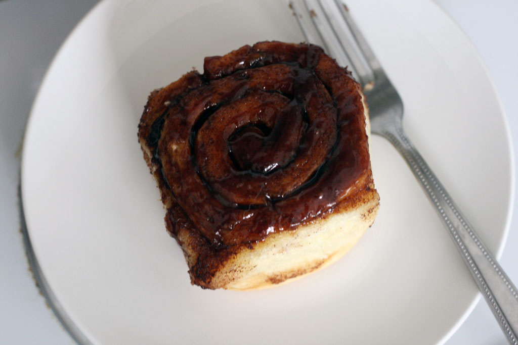 cinnamon bun on a white plate with goo dripping down and a fork to the right